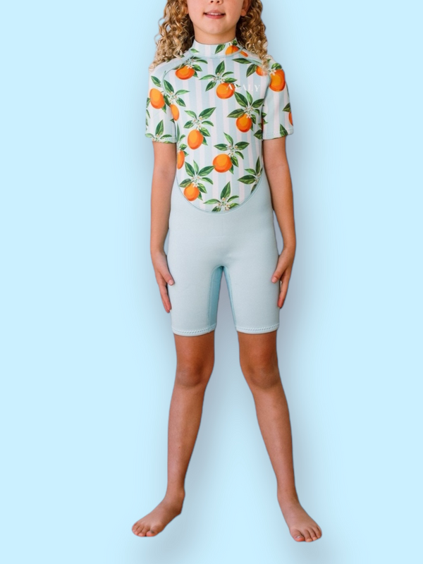 Child model wearing Blue Mandarins NOOSA wetsuit with orange citrus print and light blue shorts against blue background