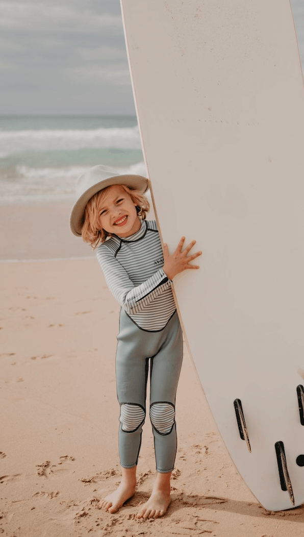 Happy child in striped wetsuit and hat waving while standing next to surfboard on beach
