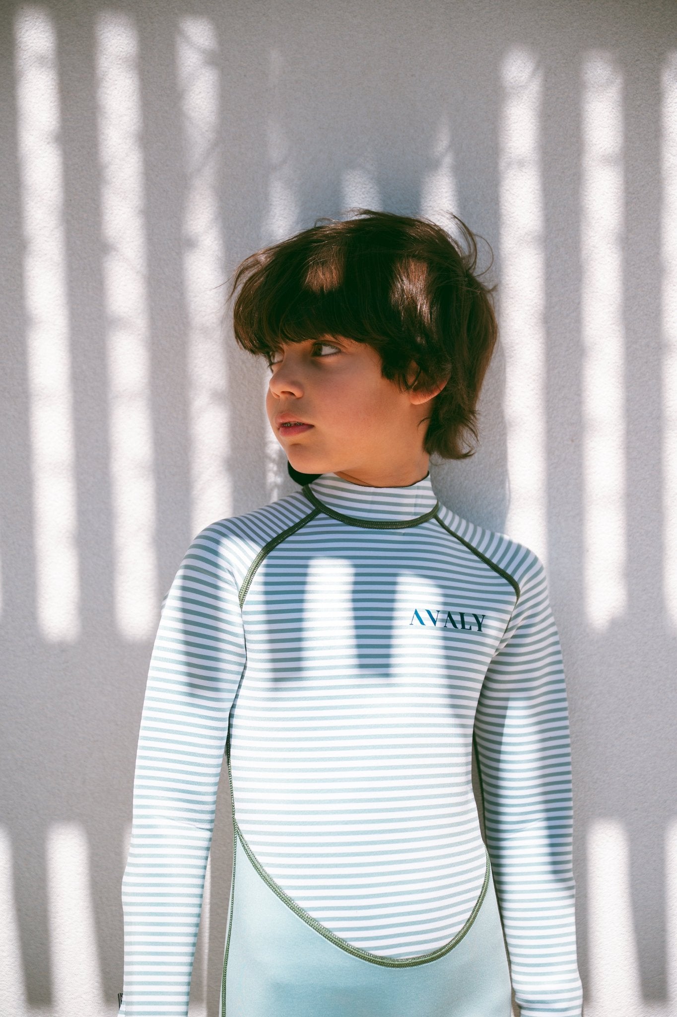 Child wearing blue and white striped Avaly wetsuit with dramatic window shadow patterns on wall behind them.