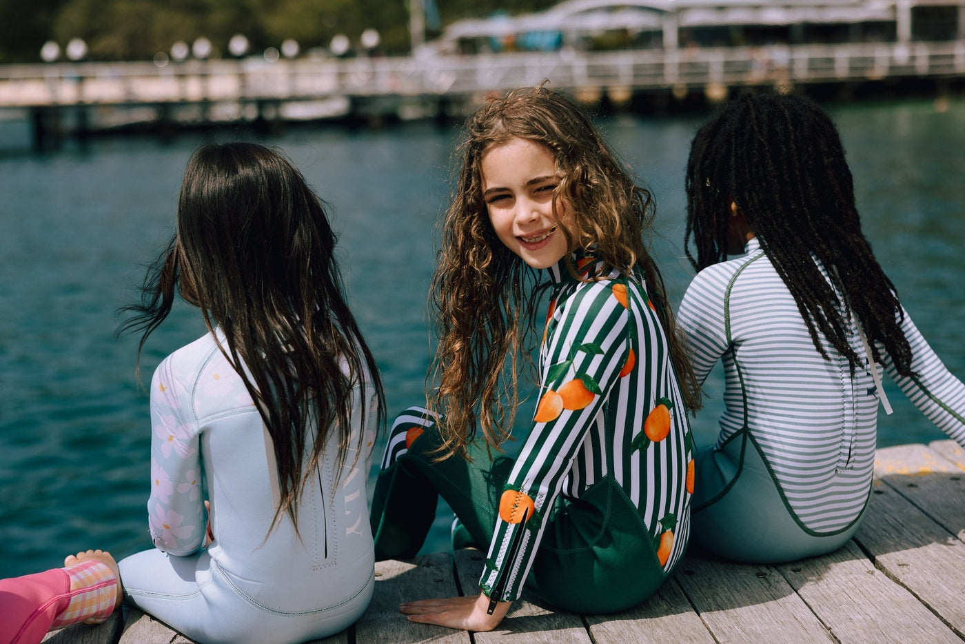 Three children in colorful wetsuits sitting on a wooden dock by water, with one girl smiling at the camera