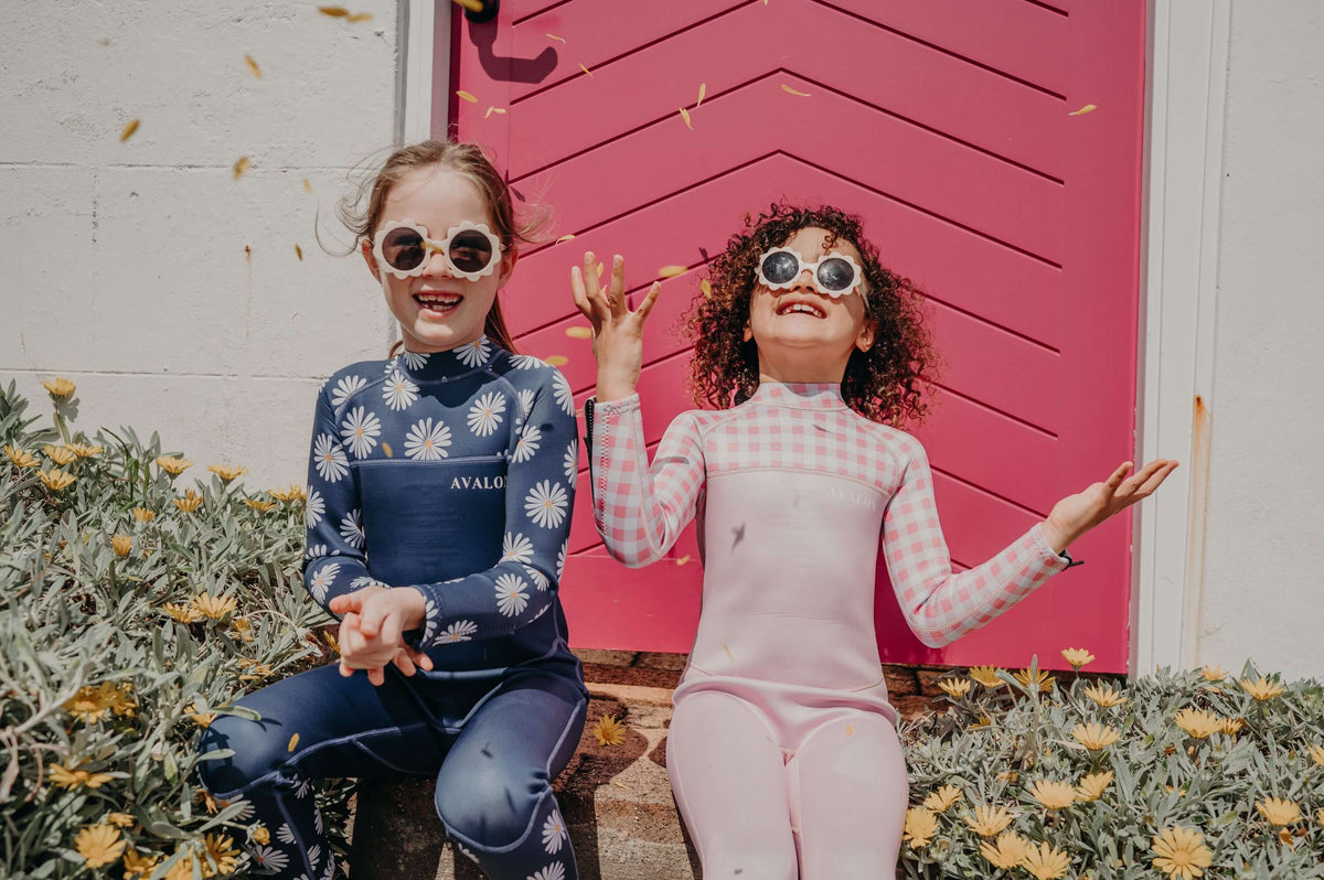 Two girls in colorful Avaly swimwear and sunglasses sitting outdoors, one waving at the camera with yellow flowers nearby