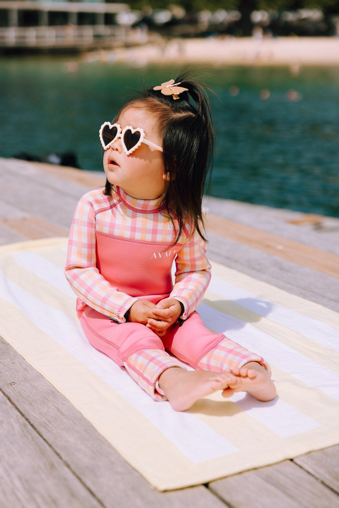 Young girl sitting poolside wearing Pink & Orange Gingham sustainable wetsuit with heart shaped sunglasses