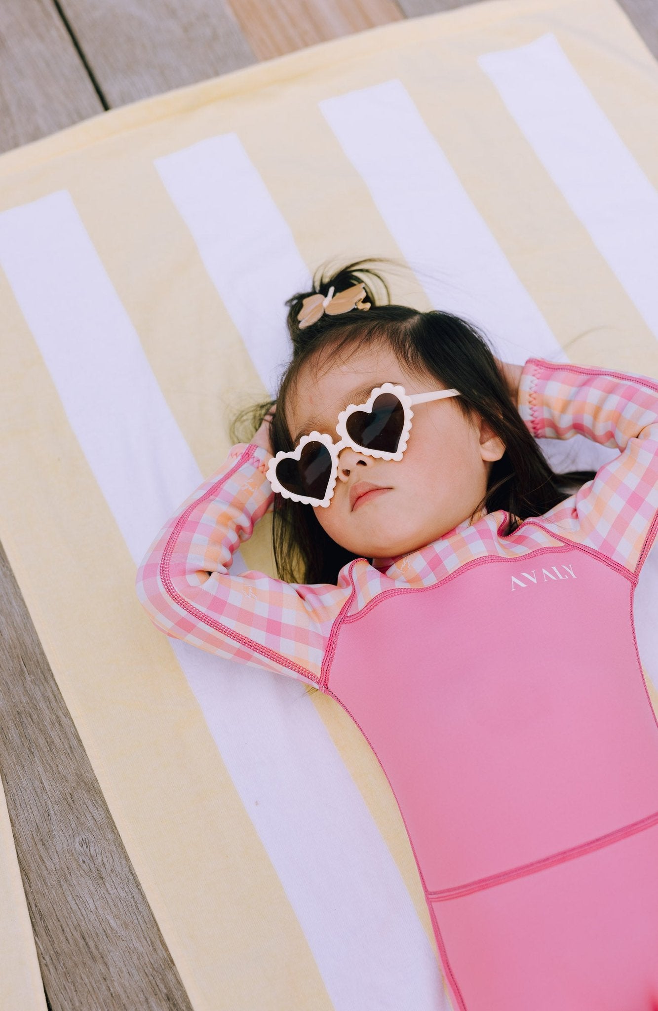 Young girl relaxing in Pink & Orange Gingham wetsuit with heart shaped sunglasses on striped towel