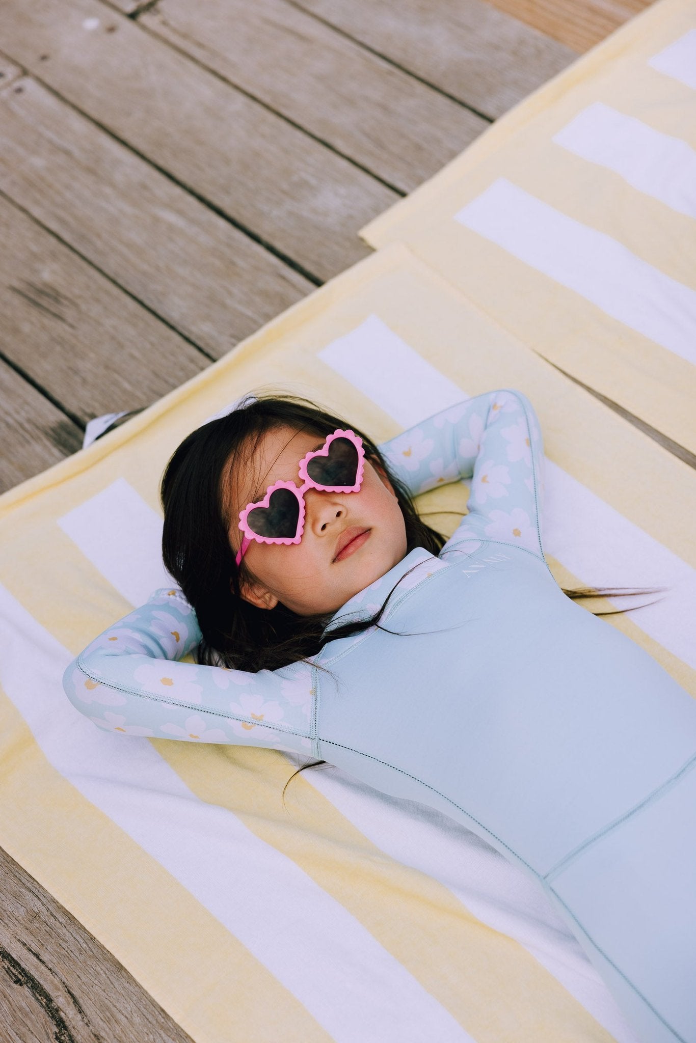 Child relaxing in mint green Mint Daisy wetsuit with small floral print, wearing heart shaped sunglasses on striped towel