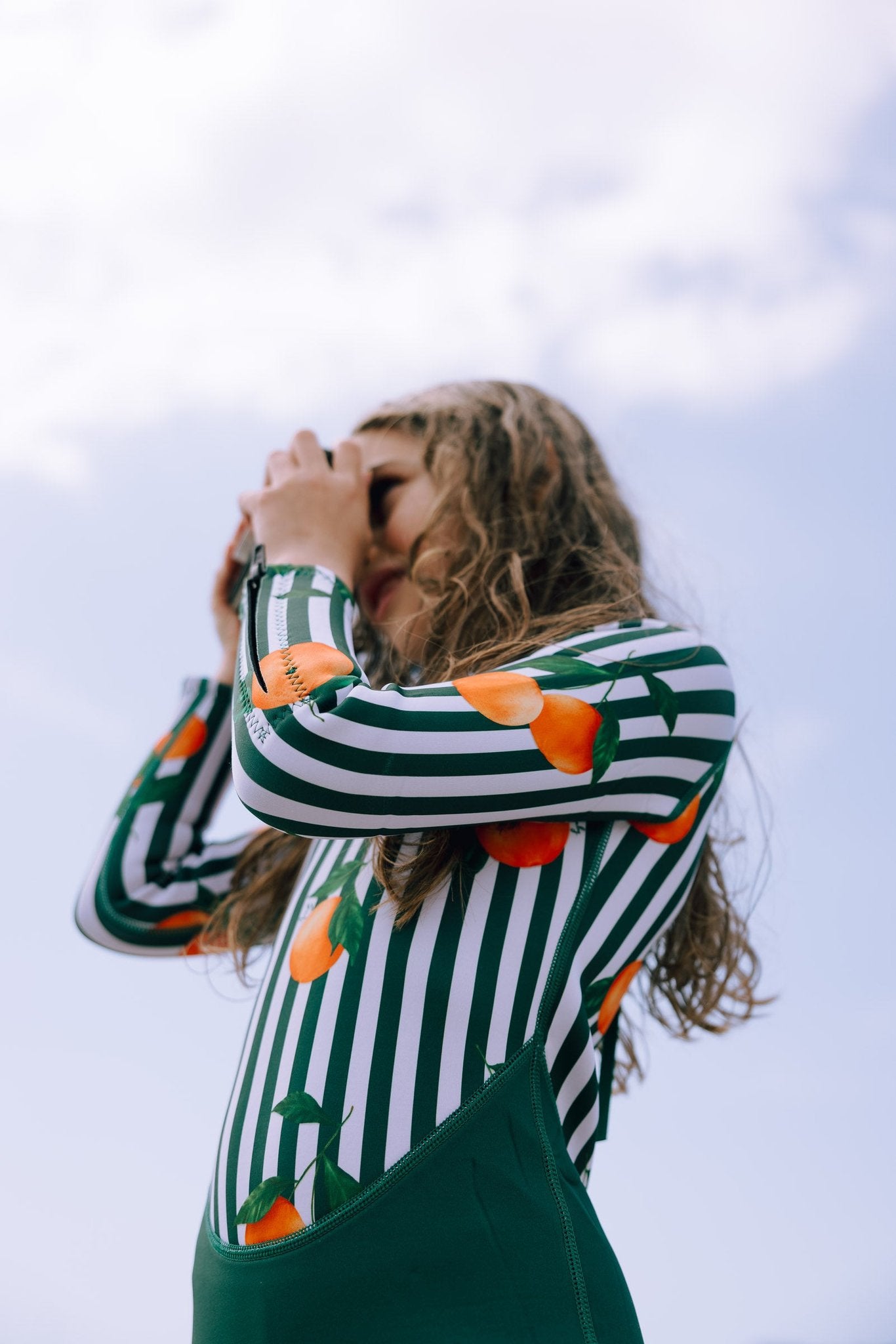 Girl in Forest Green & Mandarins wetsuit with orange citrus print on green and white stripes, hands in hair against sky