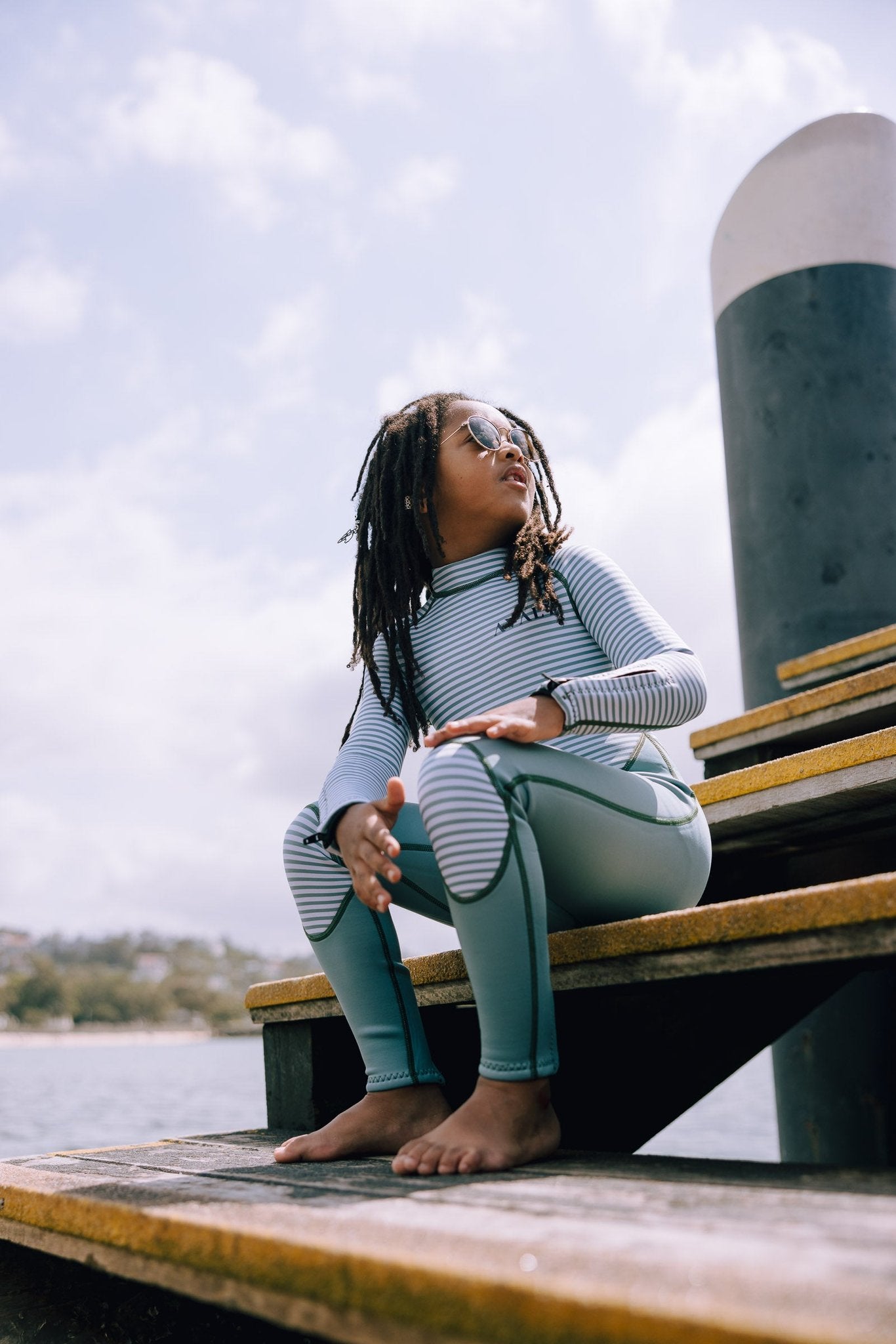 Child model in Green Striped Kids Sustainable Wetsuit sitting on wooden dock, showing striped top and green leggings