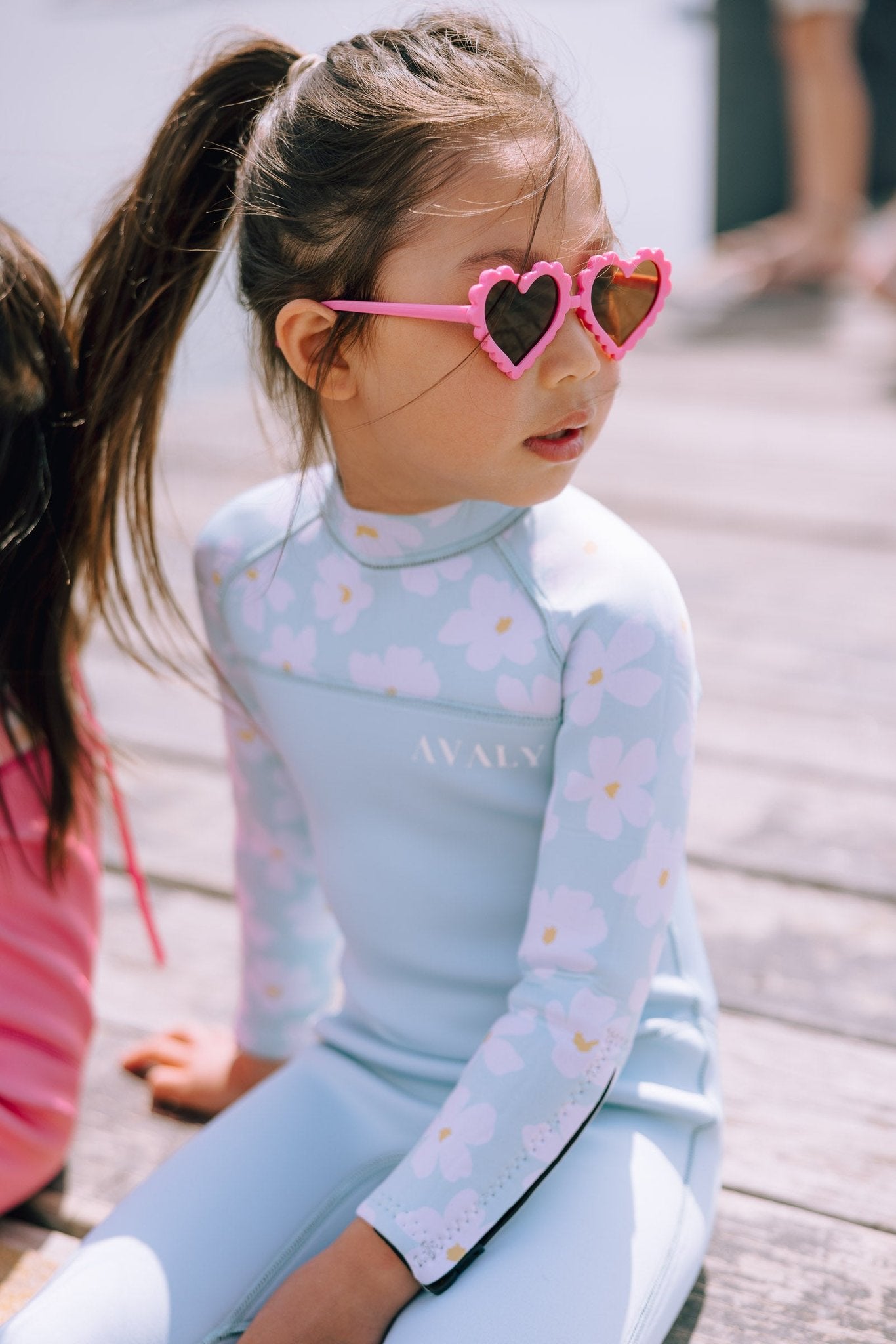 Child wearing Mint Daisy wetsuit with pink heart sunglasses, showing light blue base with daisy print pattern
