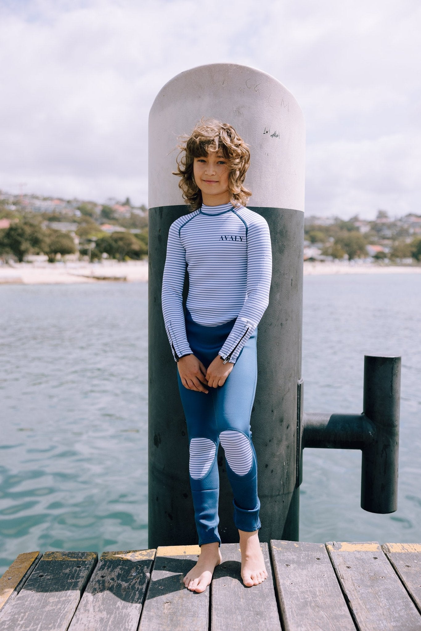 Child in Navy & White Striped wetsuit with knee patches standing on wooden dock by water