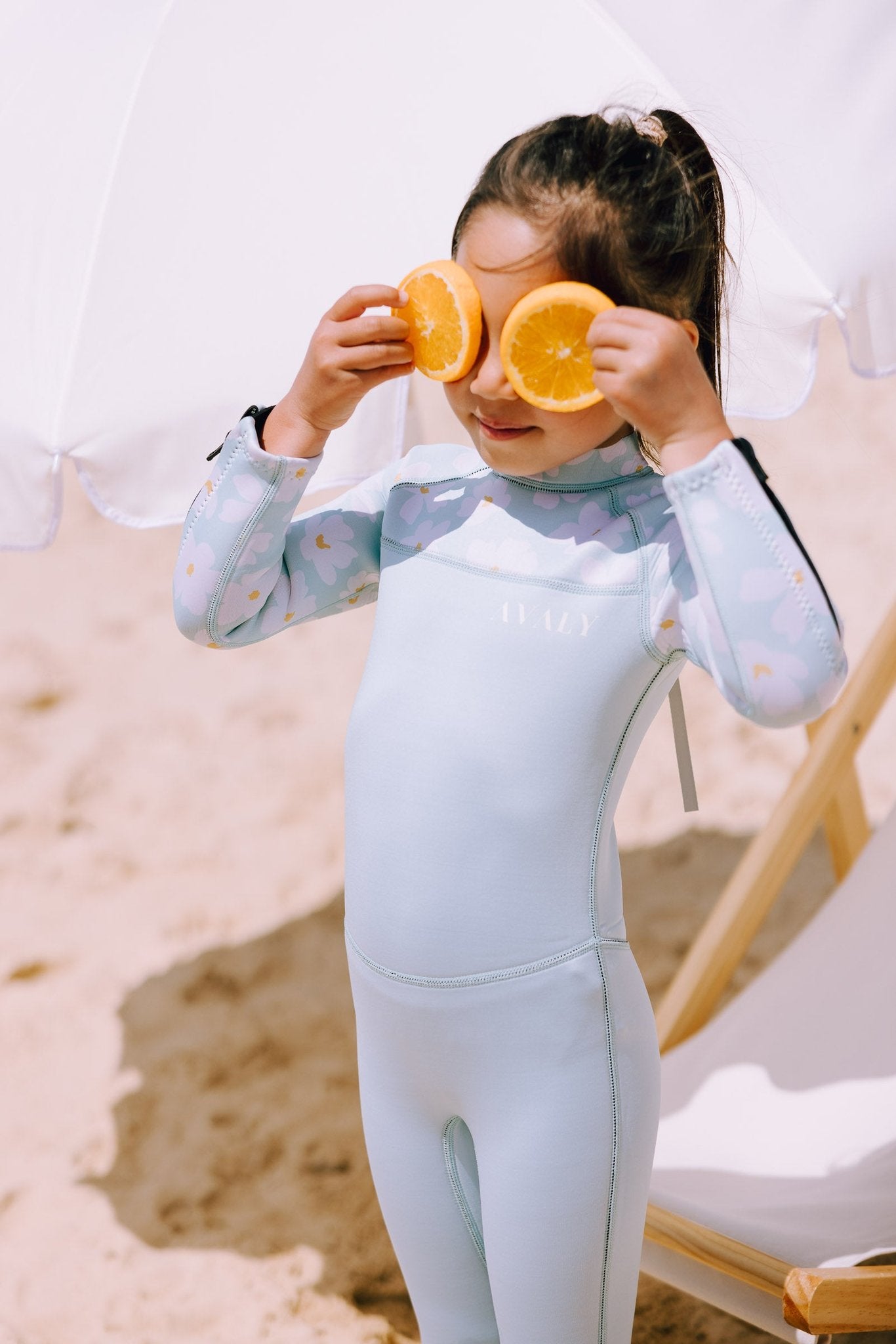Girl holding orange slices over eyes wearing mint Daisy Kids Sustainable Wetsuit with daisy print sleeves