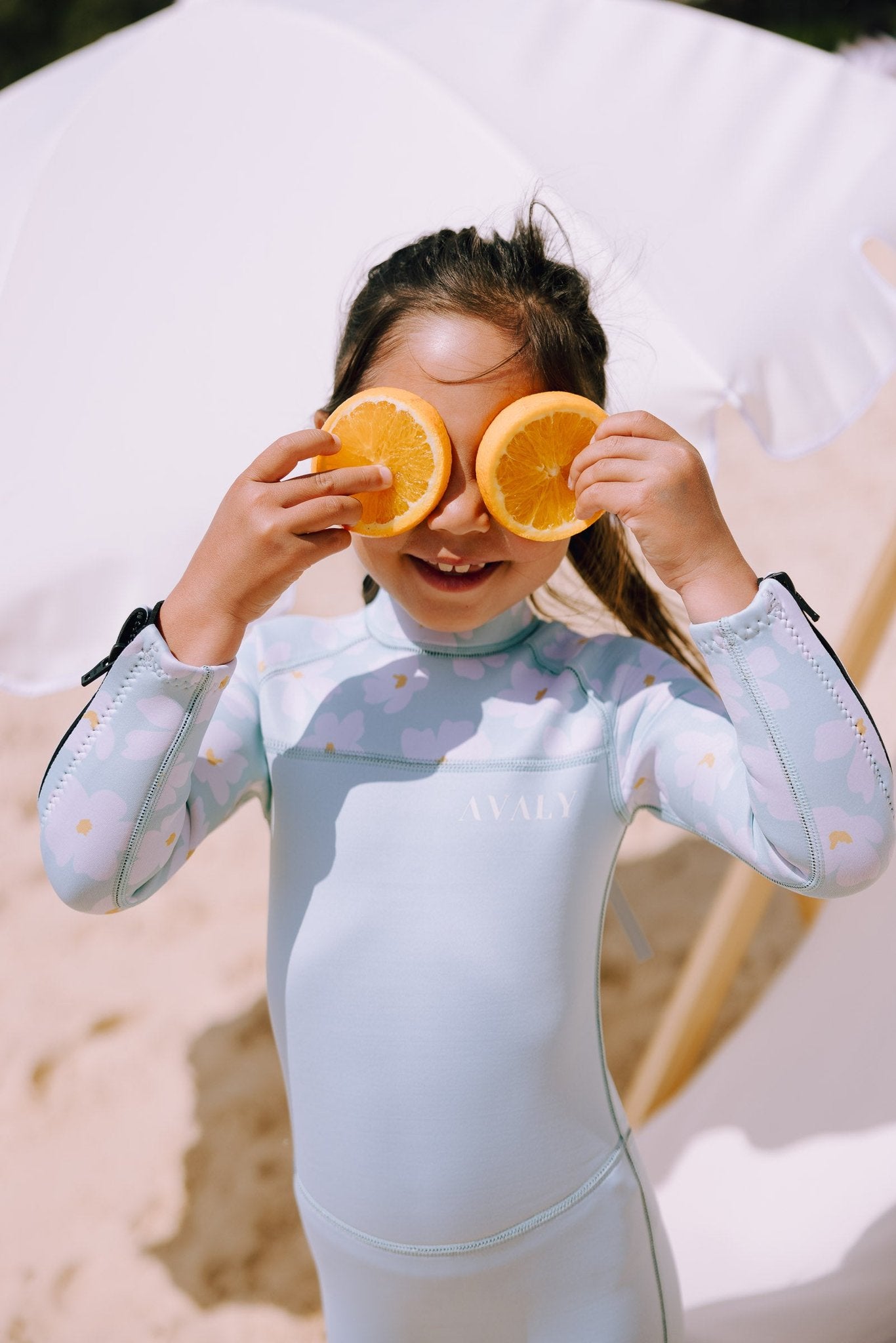 Child wearing Mint Daisy wetsuit with cloud and daisy print, playfully holding orange slices over eyes on beach