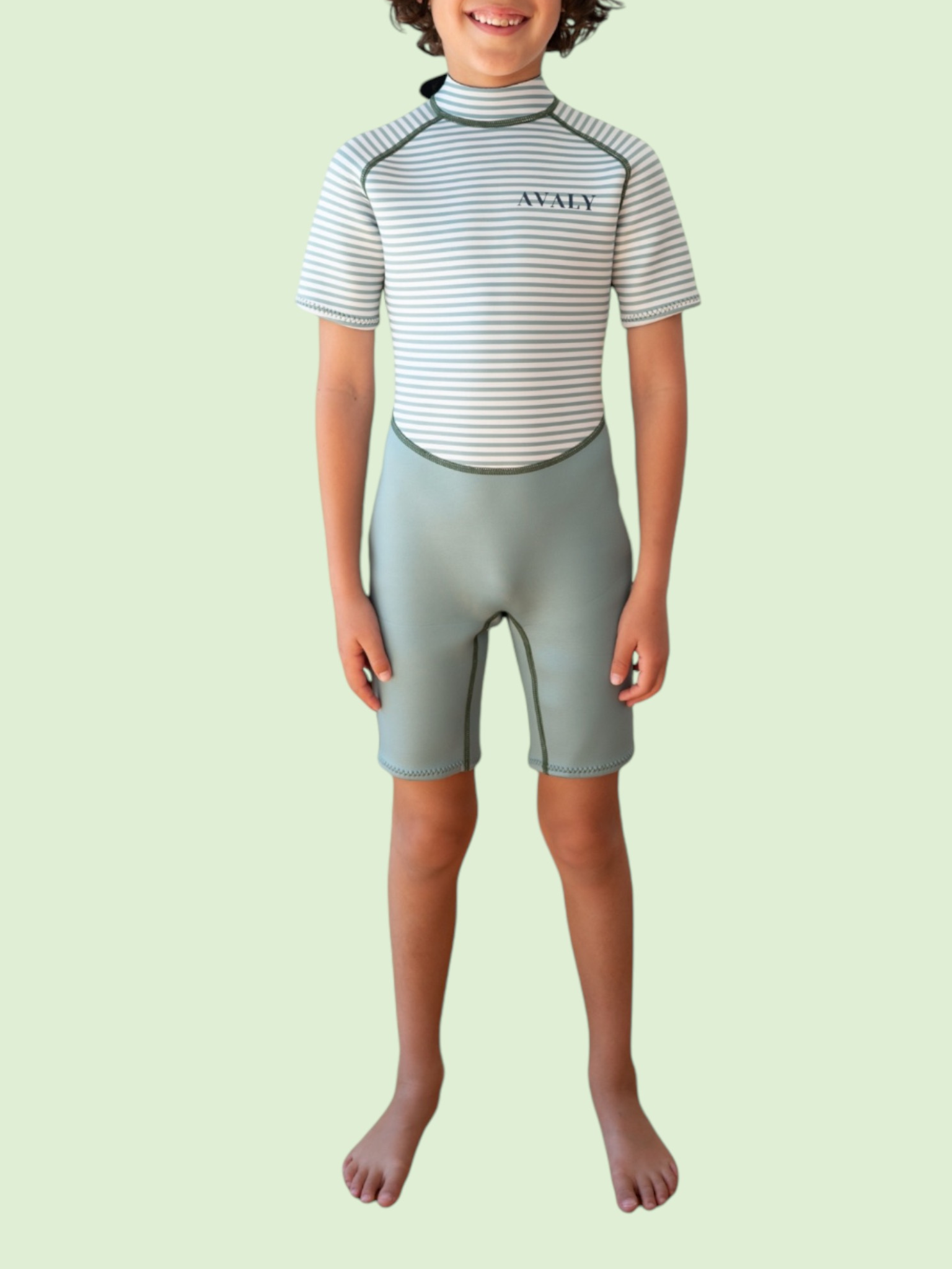 Child wearing Green Stripe Kids Wetsuit with striped short sleeve top and sage green shorts, standing against light background