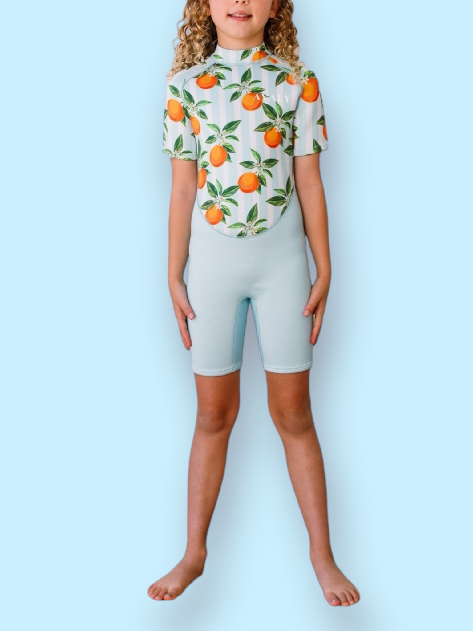 Child model wearing Blue Mandarins NOOSA wetsuit with orange citrus print and light blue shorts against blue background