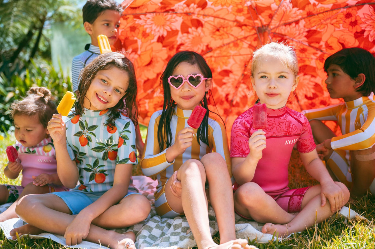 Kids in AVALY swimwear eating icy poles under an orange umbrella