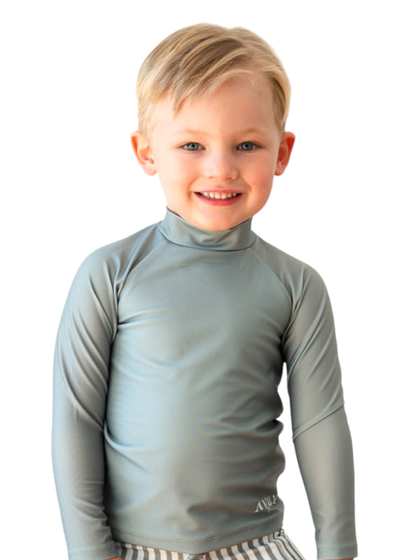 Young boy wearing sage green Manly Sustainable Rash Top with long sleeves and high neck collar, smiling at camera