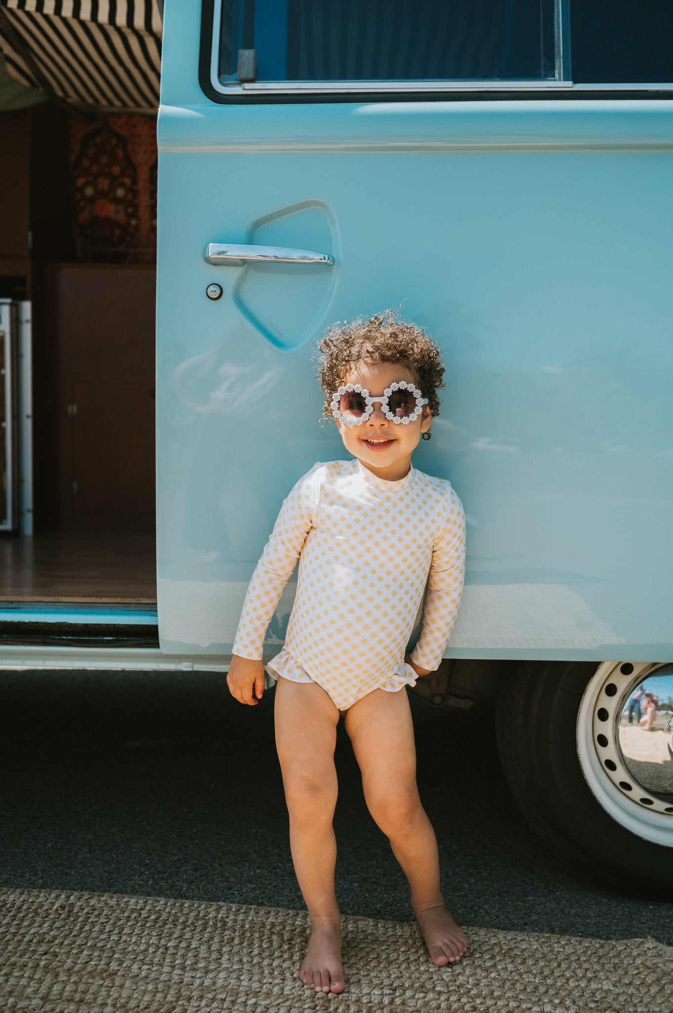 Girl in Yellow Gingham Noosa Long Sleeve One Piece with white checkered pattern standing beside vintage blue van