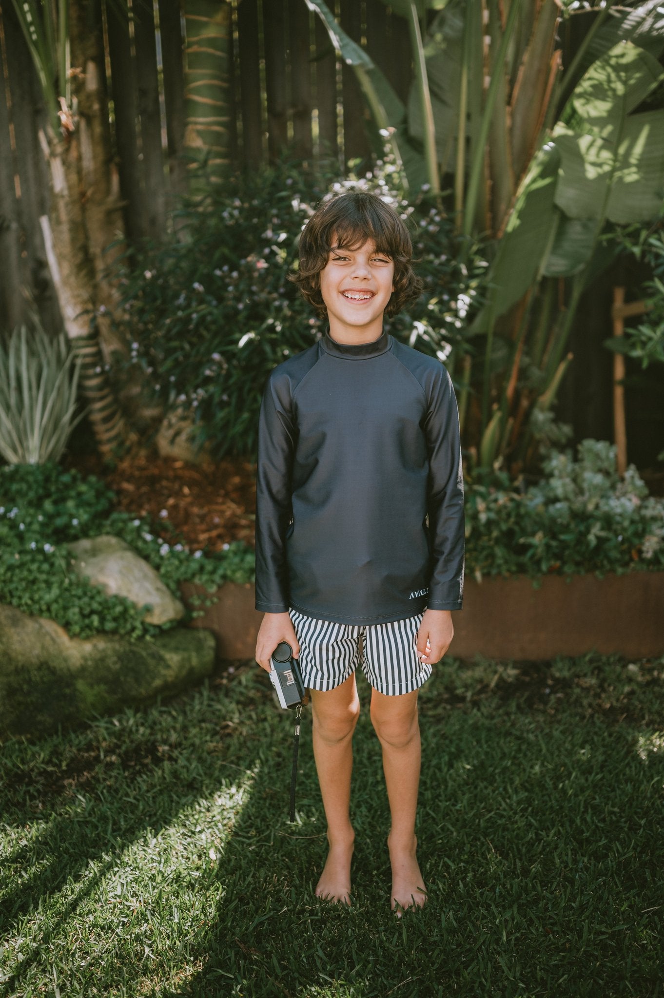 Boy wearing Navy Rash Top Byron with striped swim shorts, standing barefoot in tropical garden setting