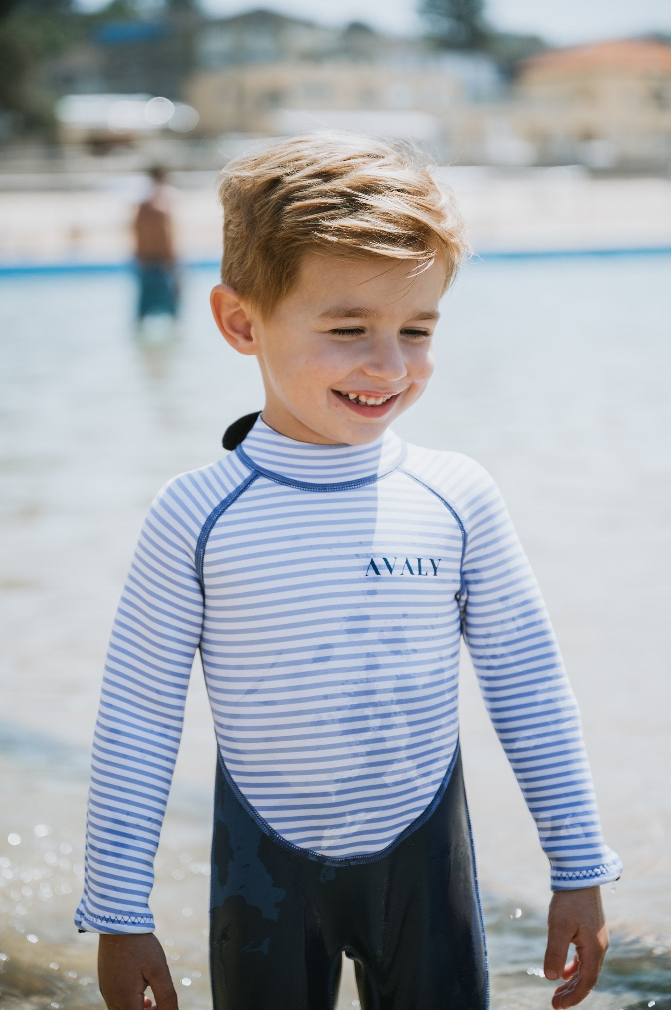 Smiling boy at beach wearing Navy & White Striped Kids Sustainable Wetsuit with blue and white horizontal stripes