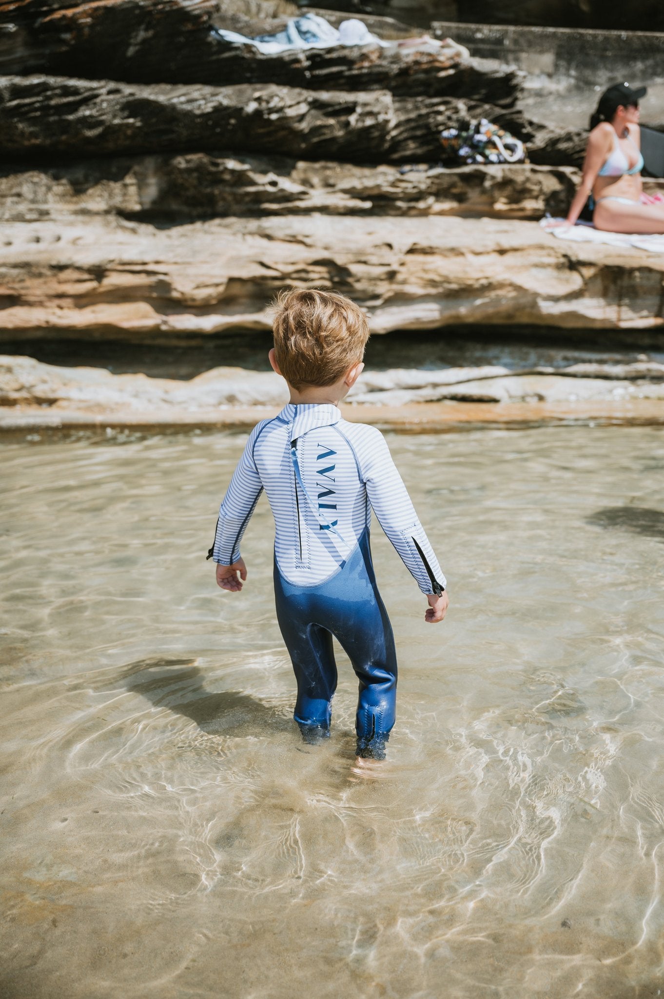 Child wearing Navy & White Striped Kids Sustainable Wetsuit wading in shallow ocean water at rocky beach