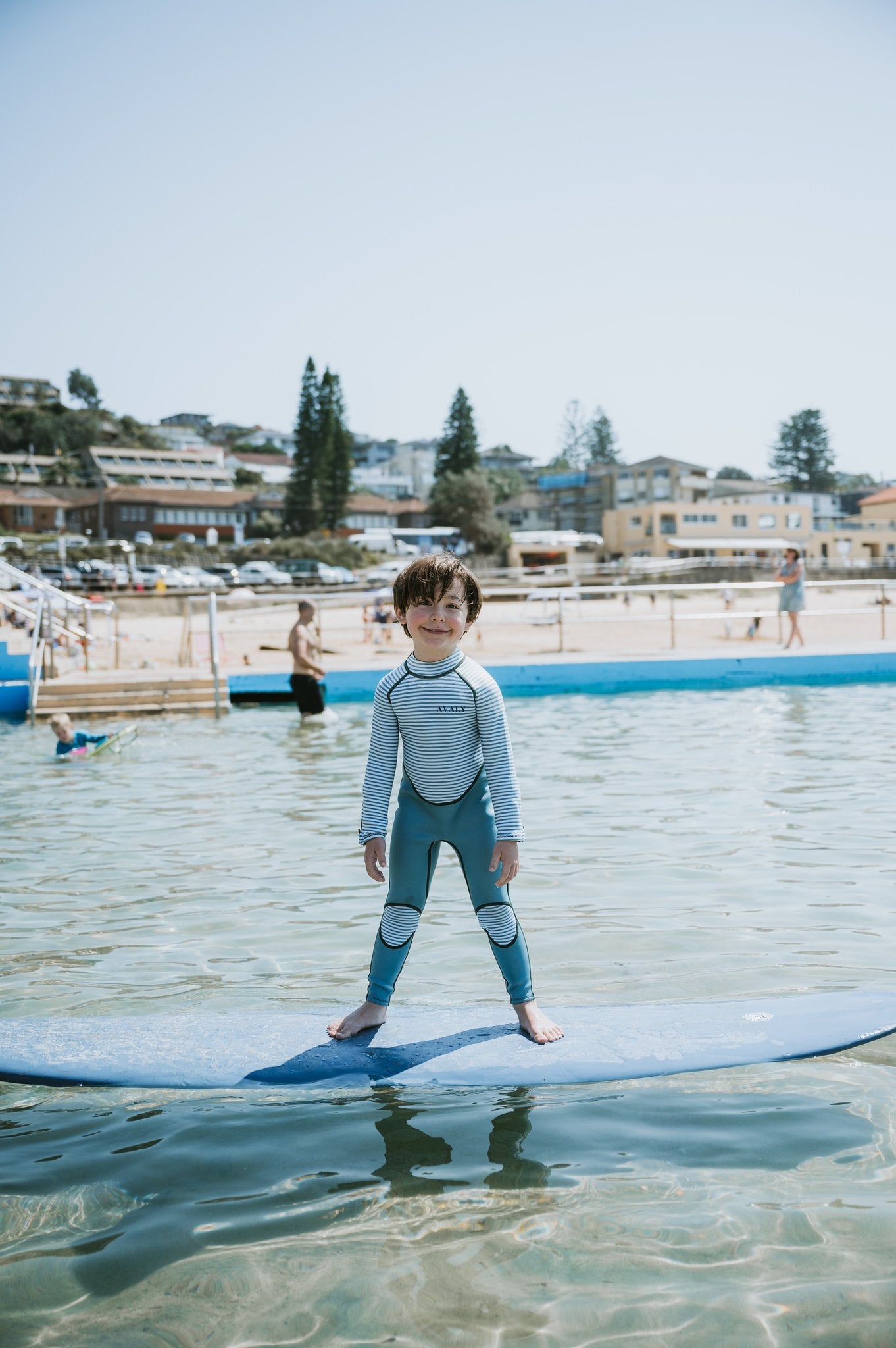 Child standing on surfboard in water wearing Green Striped Kids Sustainable Wetsuit with blue and white stripes