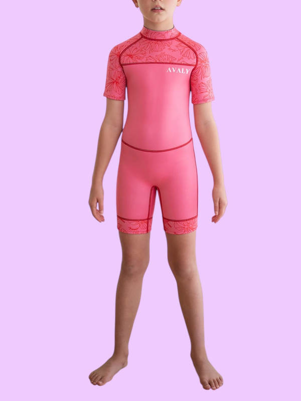 A child standing front on in the Hawaiian kids wetsuit, a sustainable pink springsuit with red hibiscus floral sleeves.