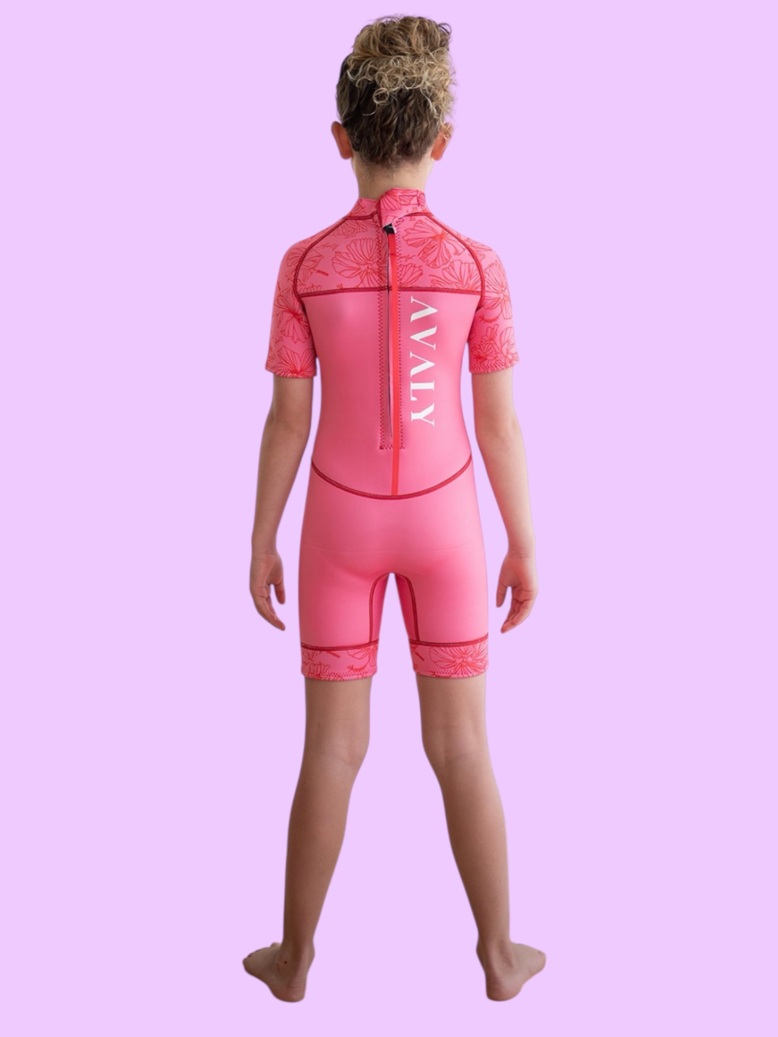 A child standing back on in the Hawaiian kids wetsuit, a sustainable pink springsuit with red hibiscus floral sleeves.